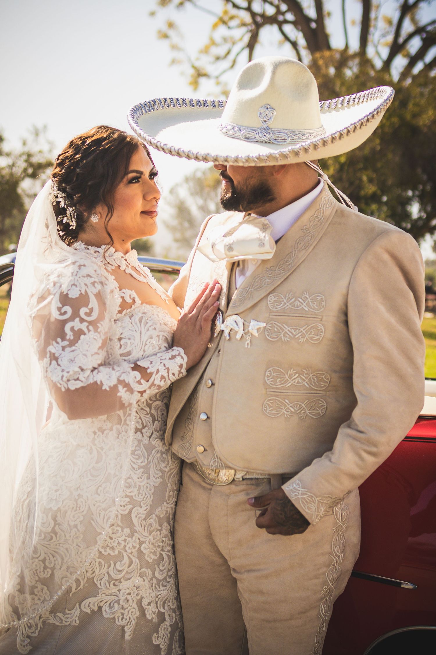 Wedding couple posing by red classic car wearing traditional Mexican wedding attire. At San Luis Rey Parish celebrating a wedding