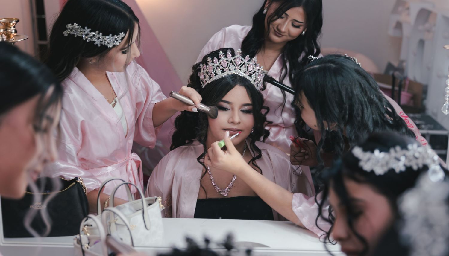 Bride in tiara getting makeup done by bridesmaids in pink robes during wedding preparation.