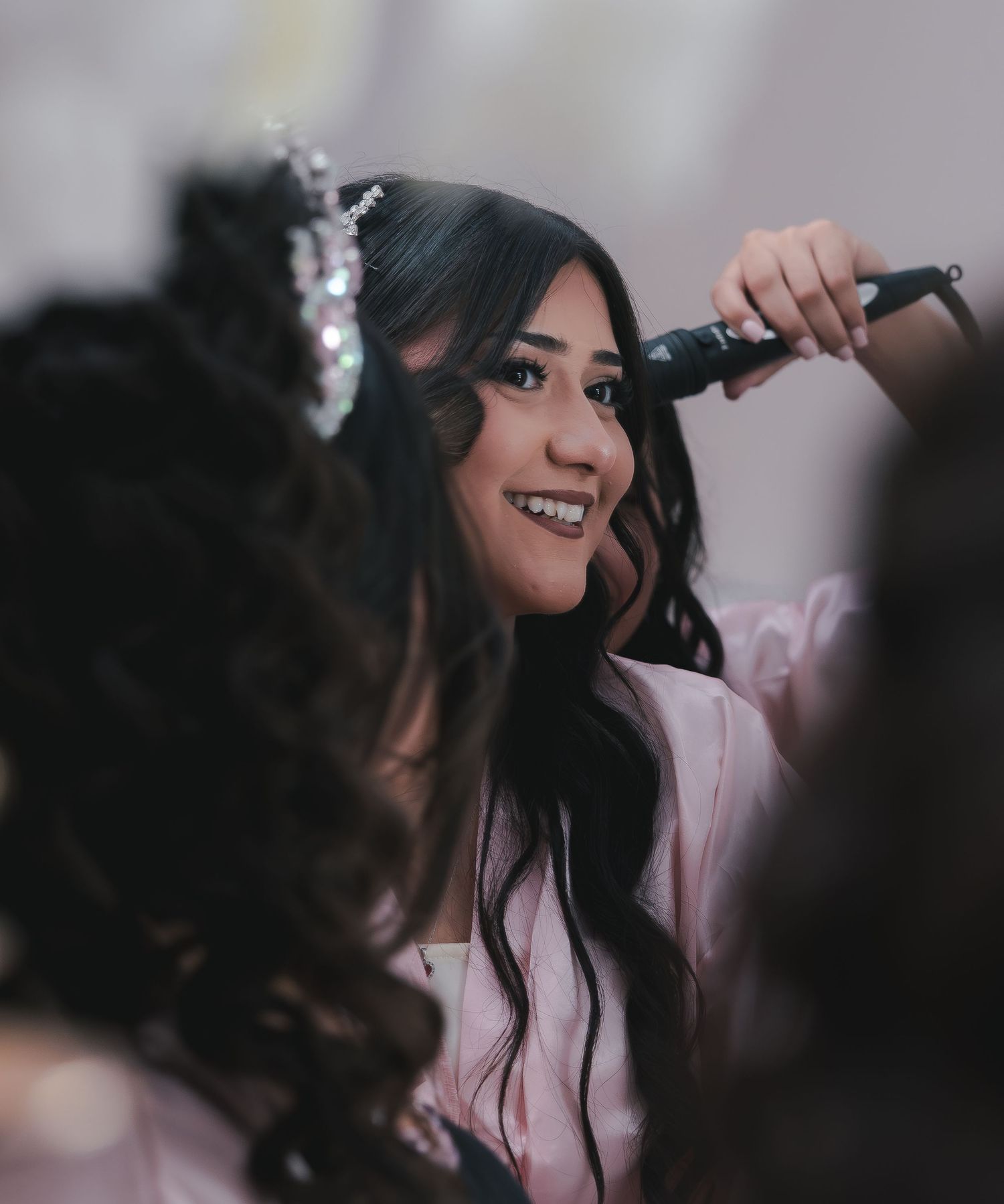 Young woman wearing a tiara getting her hair done in front of a mirror.