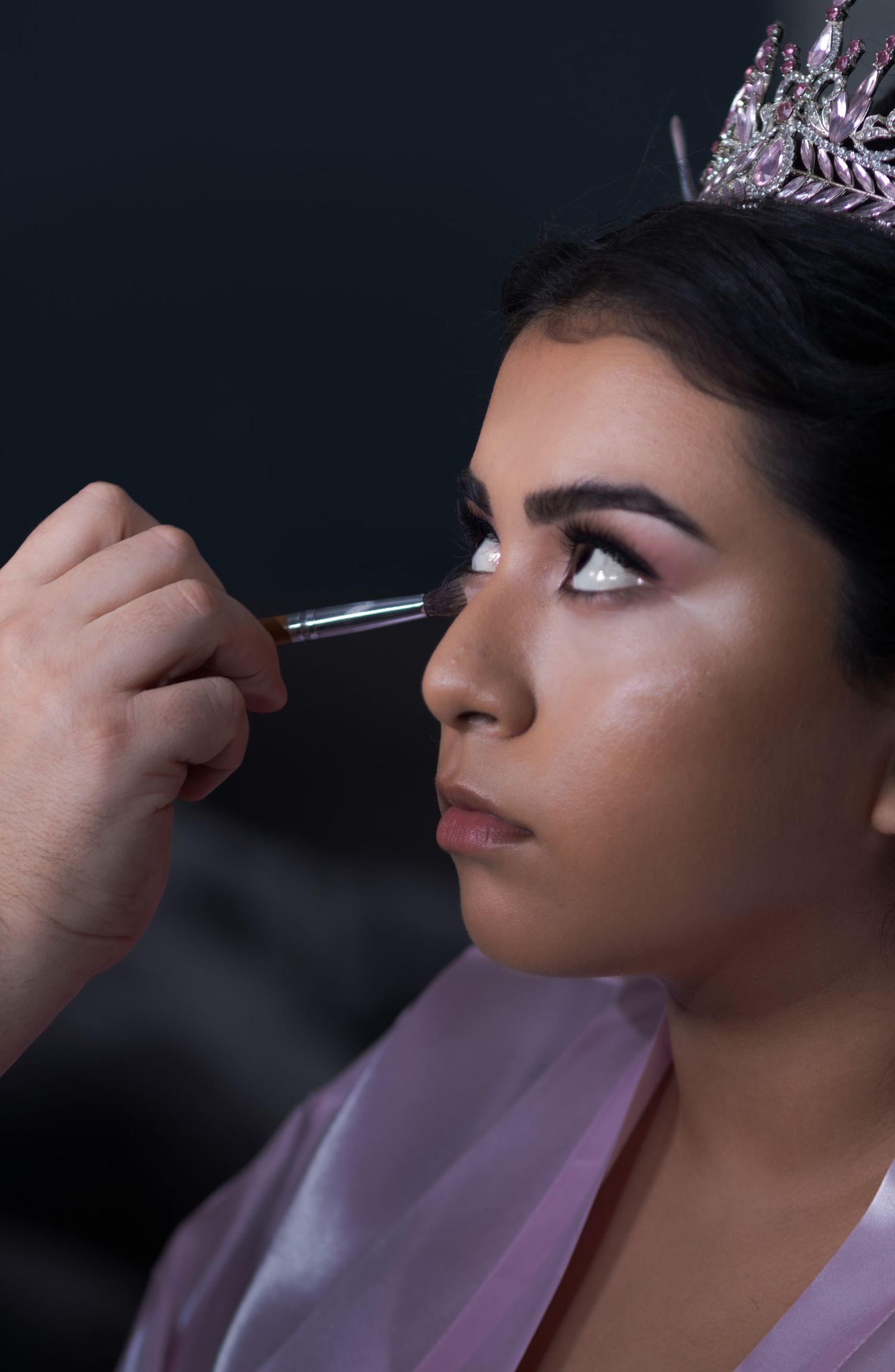 Professional makeup artist applying eye makeup with precise brush technique in dark studio setting. At a Quinceañera