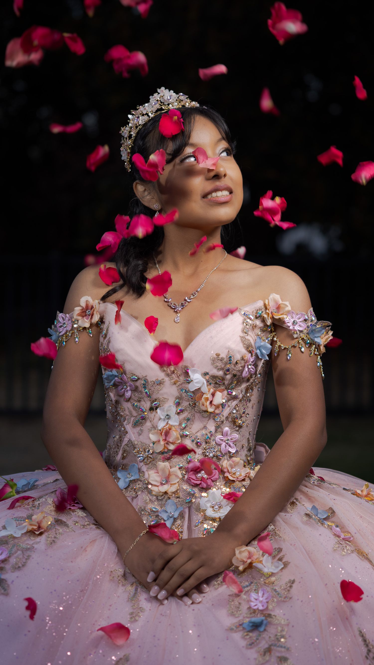 Young woman in floral quinceañera gown and tiara smiles as pink rose petals fall around her.