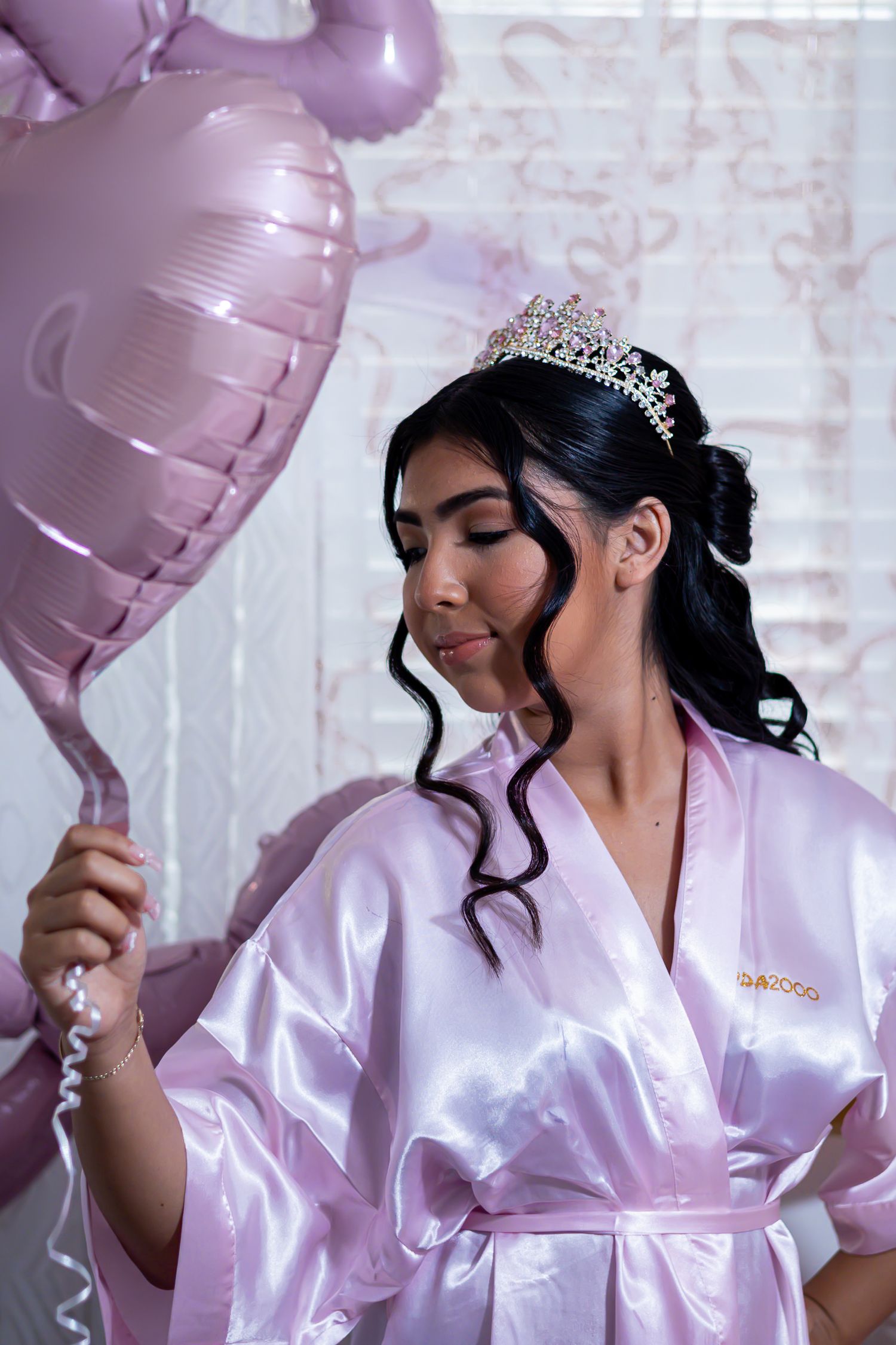 Person in pink satin robe celebrating with heart-shaped balloons against white brick wall. At a Quinceañera