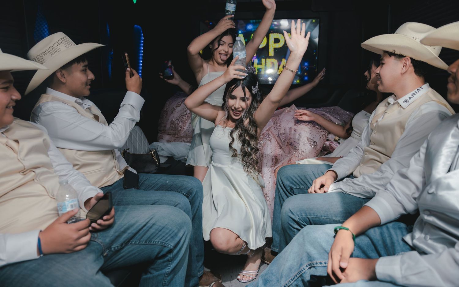 Young woman in white dress celebrates quinceañera with chambelanes in cowboy hats dancing at colorful party.