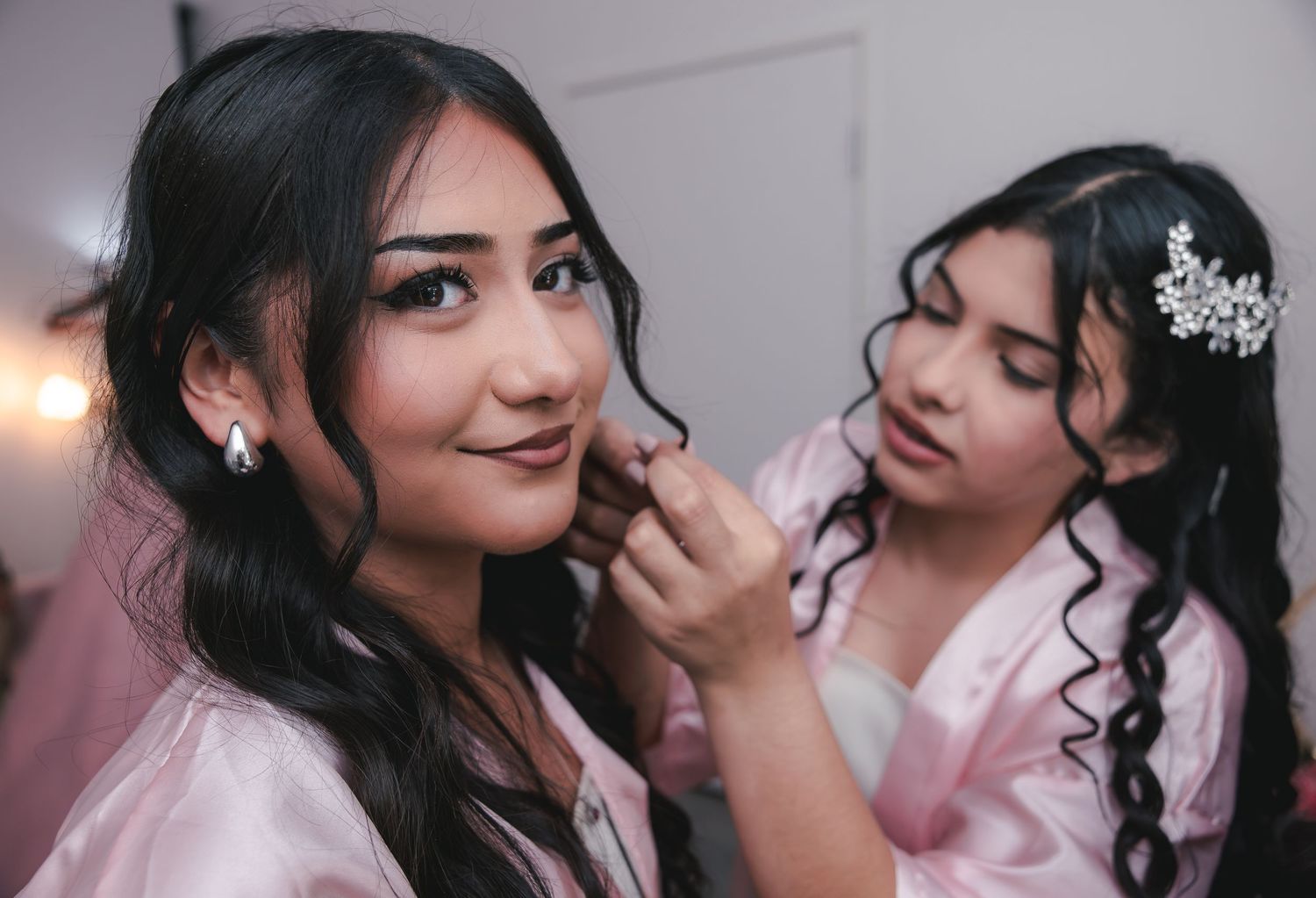 Two bridesmaids in pink robes, one applying lipstick to the other, both wearing elegant hair accessories.