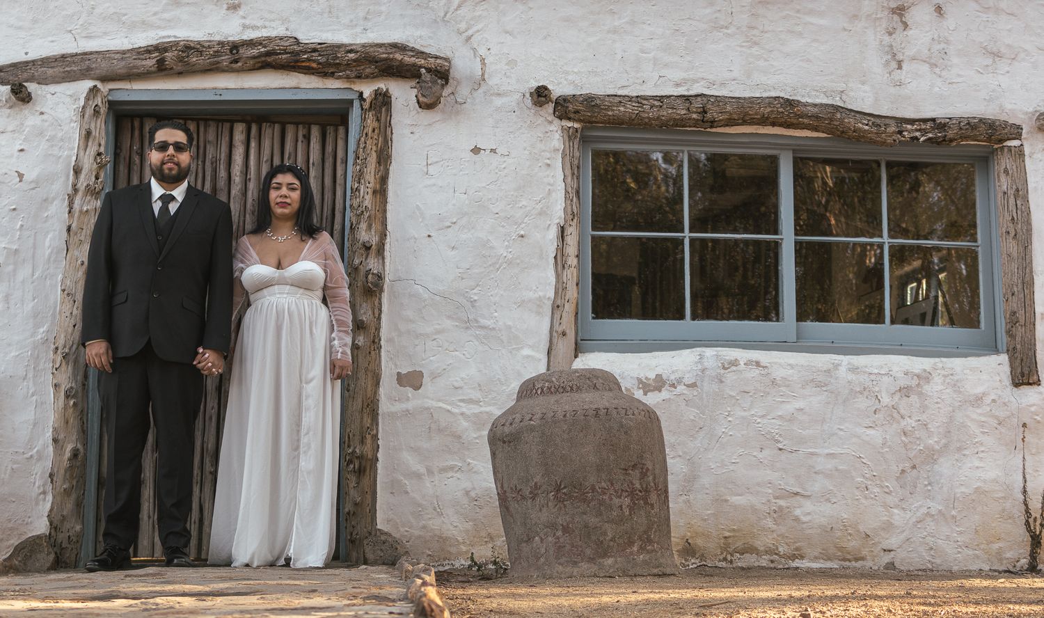 Wedding couple stands in rustic doorway of white adobe building. At Rancho Carrillo