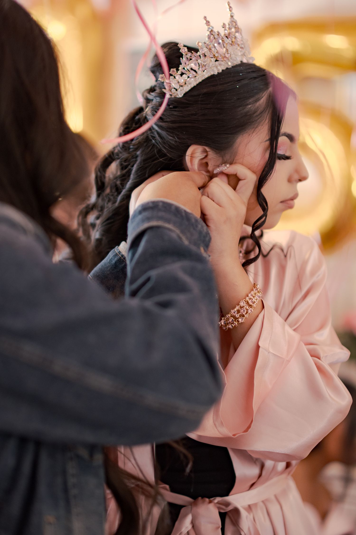 Close-up shots of a Quinceañera getting their hair and makeup done while wearing a pink robe and tiara. At a Quinceañera