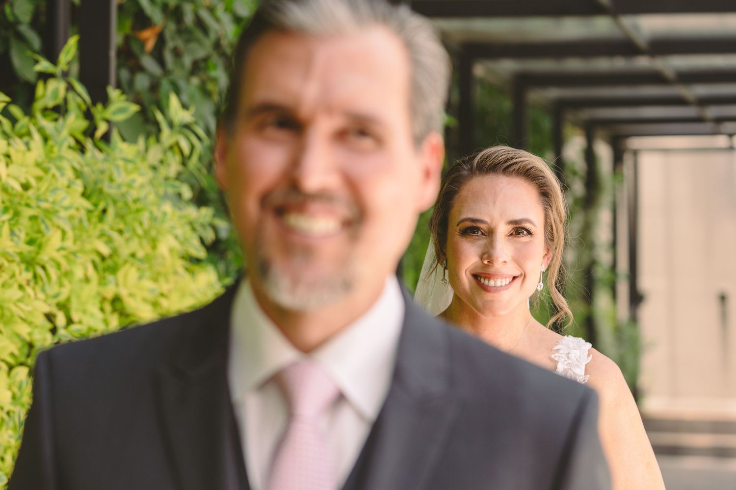 A smiling bride in a white dress stands behind a blurred man in a gray suit with pink tie at an outdoor wedding.