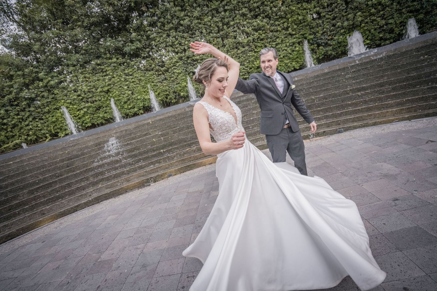 Bride in flowing white gown twirls as groom in gray suit follows during outdoor wedding photo session.