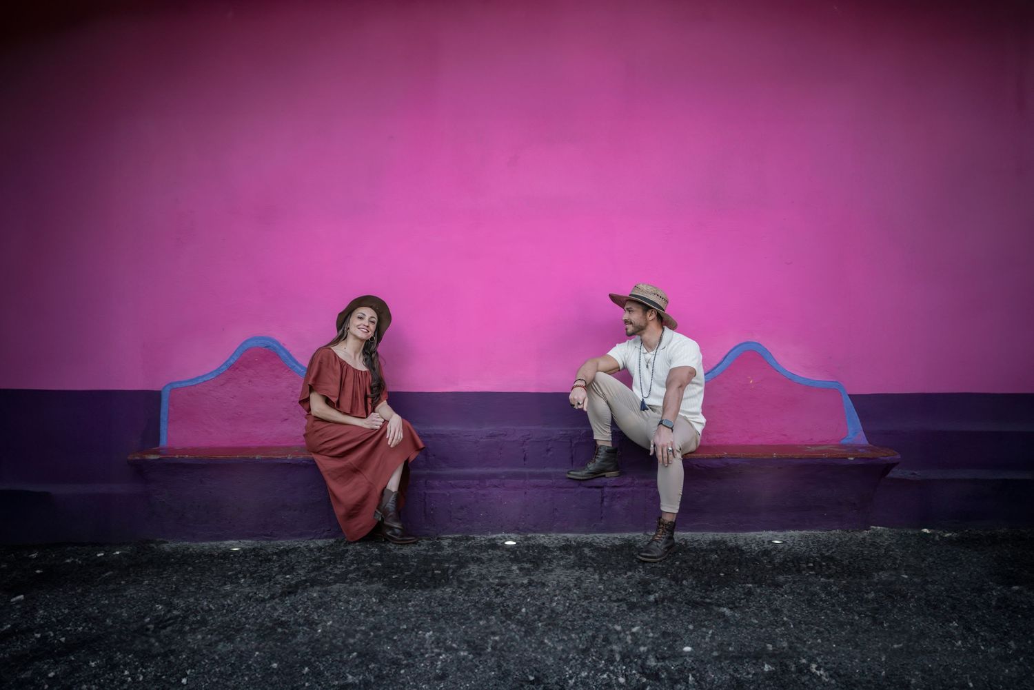 Two people sit against a pink wall with legs extended in a creative dance pose on black floor.