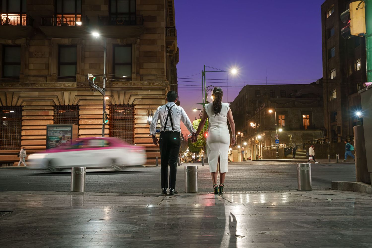 Couple holding hands on a city street corner at night with blurred passing car and glowing streetlights.
