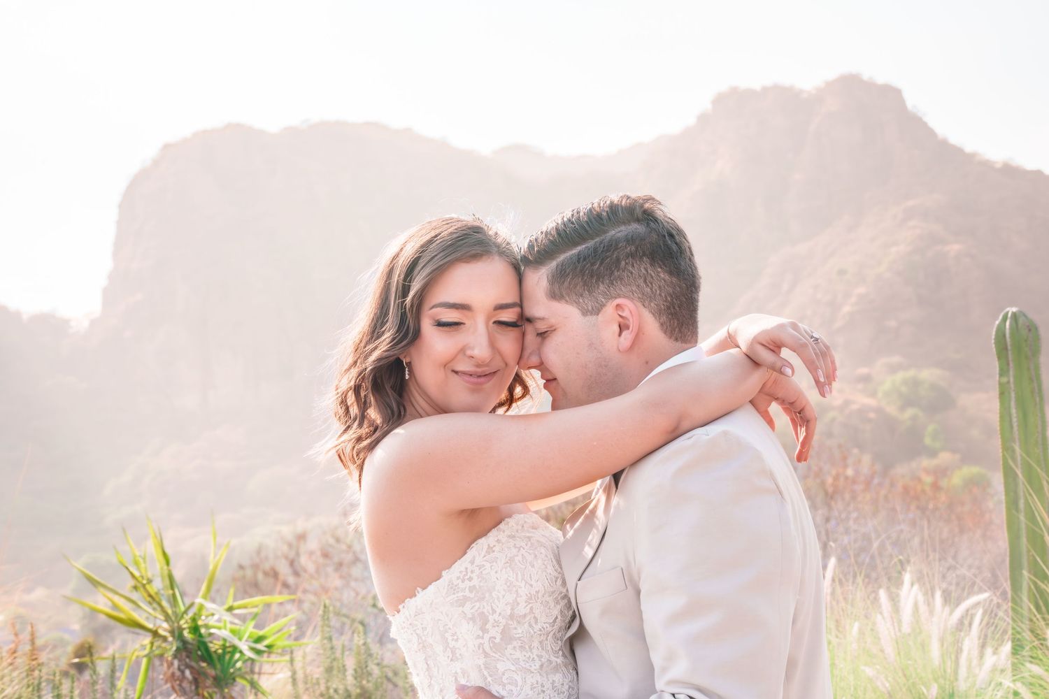 Bride and groom embrace tenderly in a sun-kissed desert landscape with mountains and cacti in the background.
