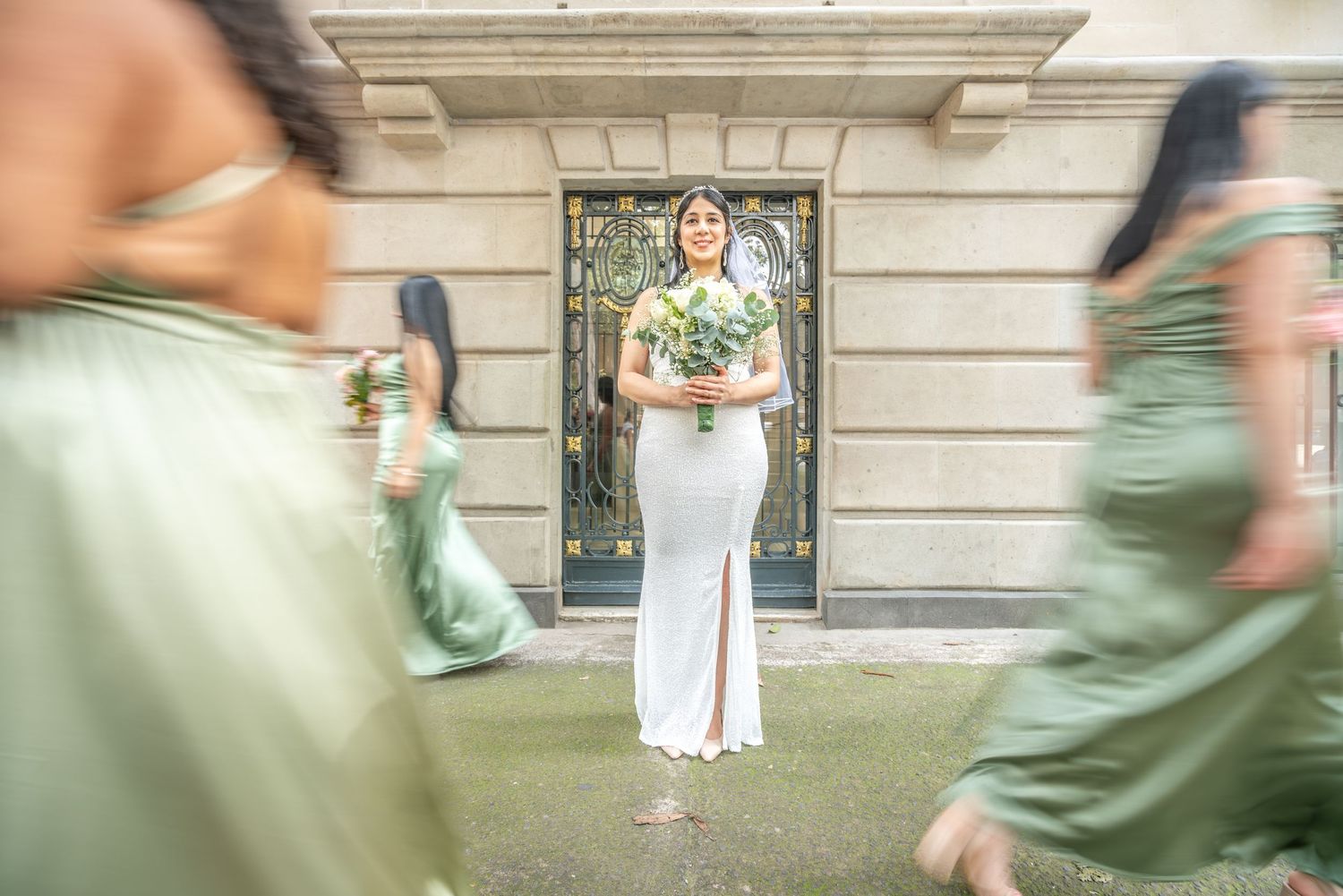 A person in white pants and green top stands in front of an ornate door while others in sage green dresses pass by.