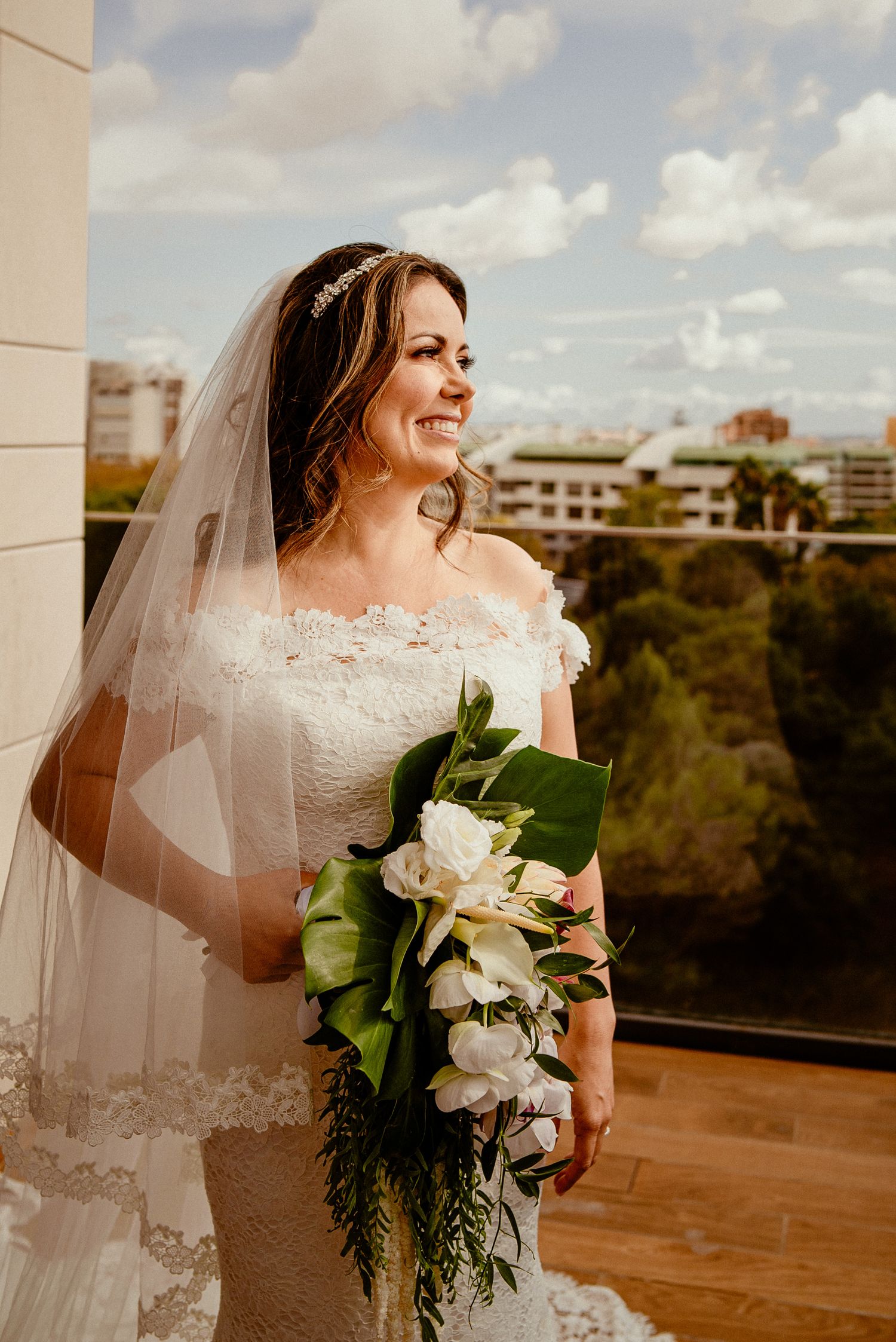 Bride in lace dress holding tropical bouquet smiling on balcony with city view.