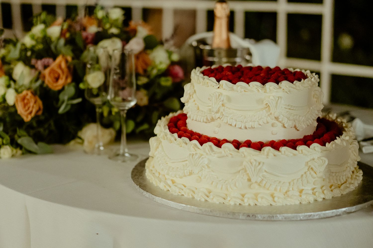 Two-tier white wedding cake with red raspberry filling and decorative cream piping on elegant white table.