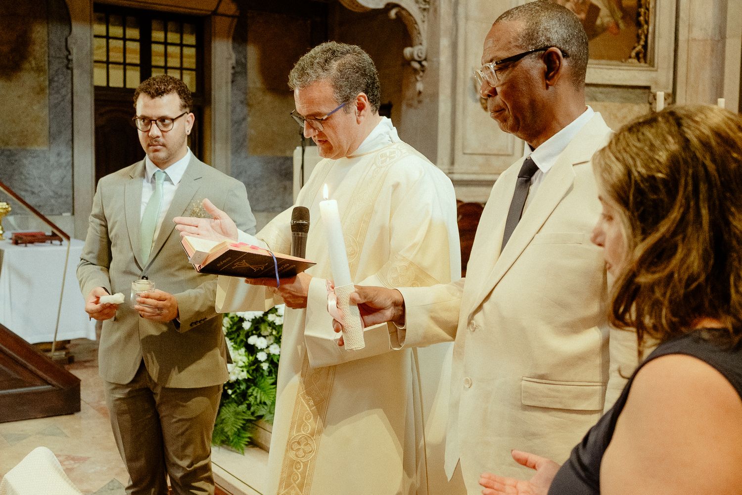 Priest in white vestments reads from a book during a church ceremony with attendees standing nearby.