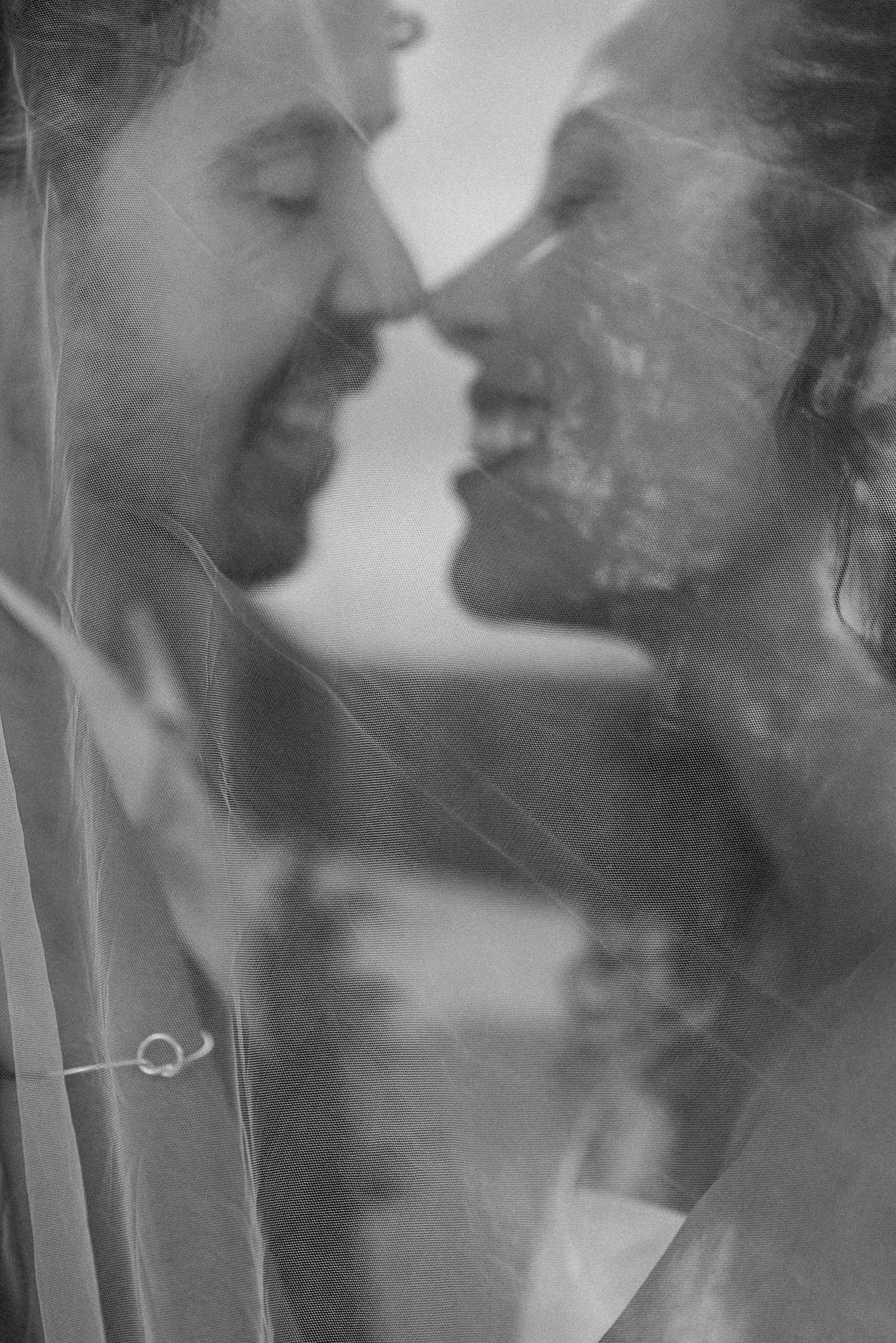 Black and white photo of a couple sharing a romantic almost-kiss beneath a wedding veil.