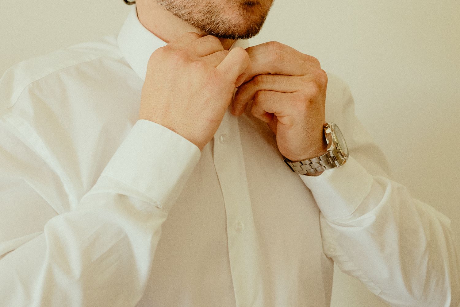 Man in white dress shirt adjusting his collar button while wearing a gold watch.