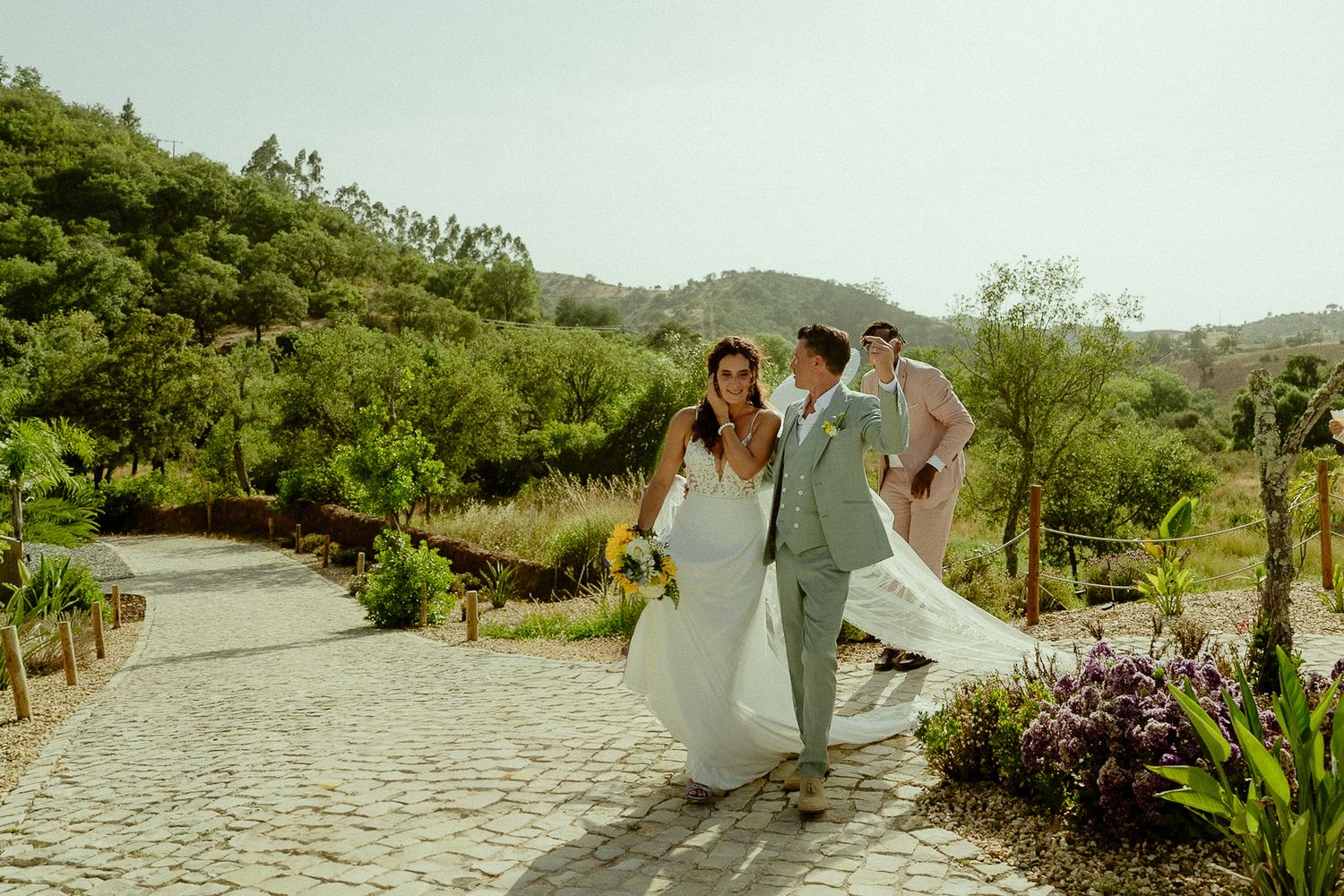 Bride and groom walk hand-in-hand on a cobblestone path surrounded by lush greenery on their wedding day.