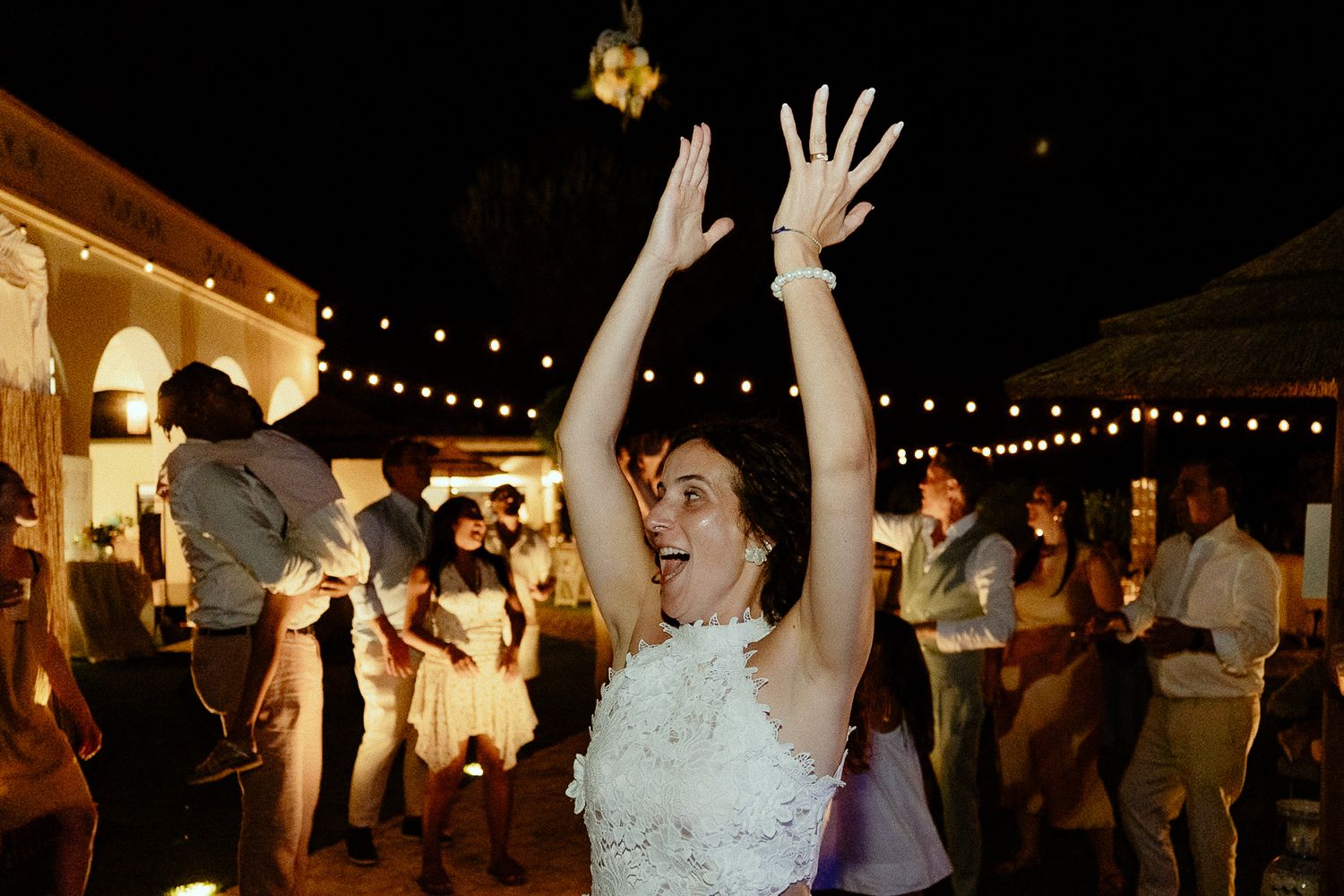 Bride in white dress dancing joyfully with arms raised at outdoor wedding reception with string lights.
