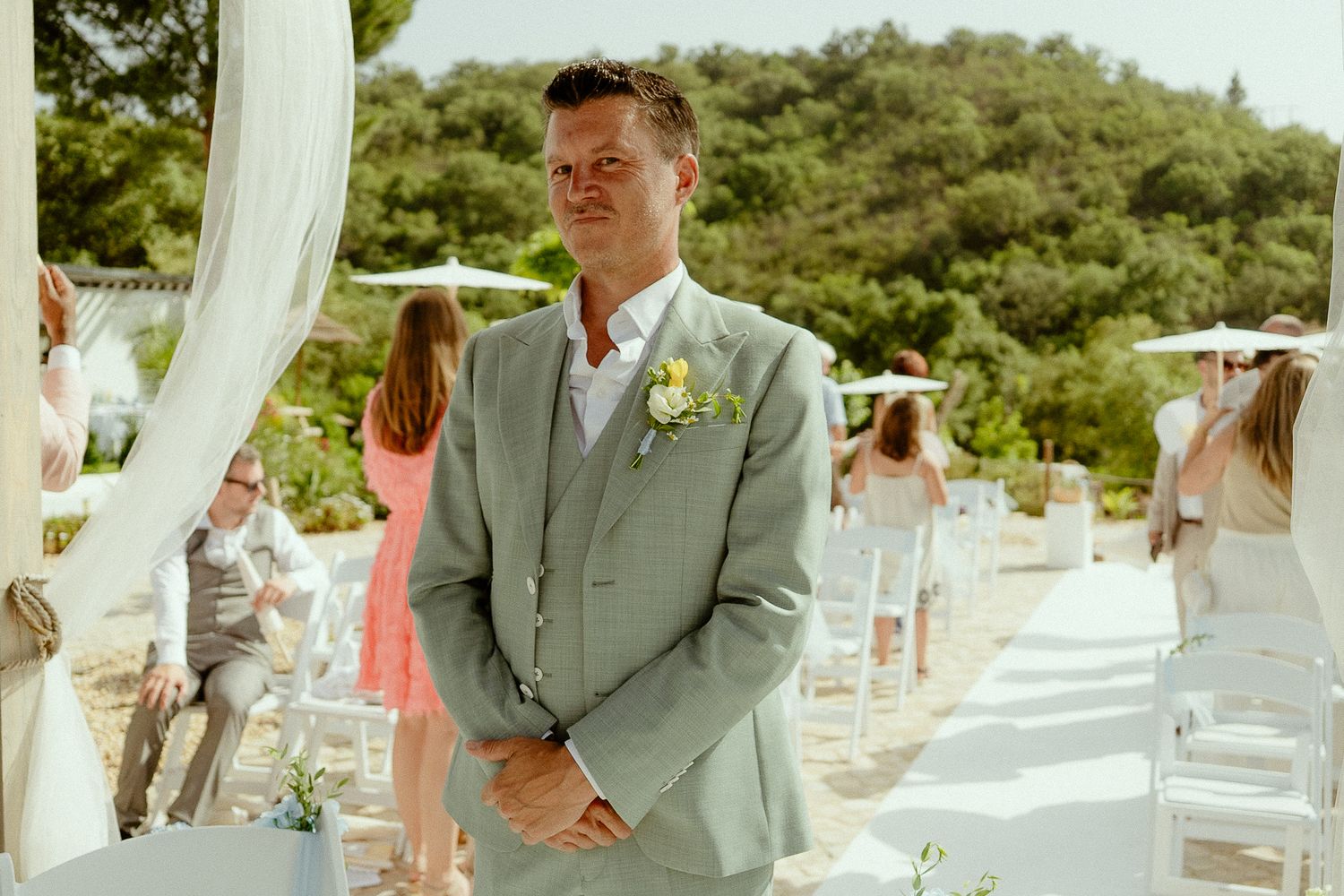 Groom in light gray suit with boutonniere stands at outdoor wedding ceremony aisle lined with white chairs.