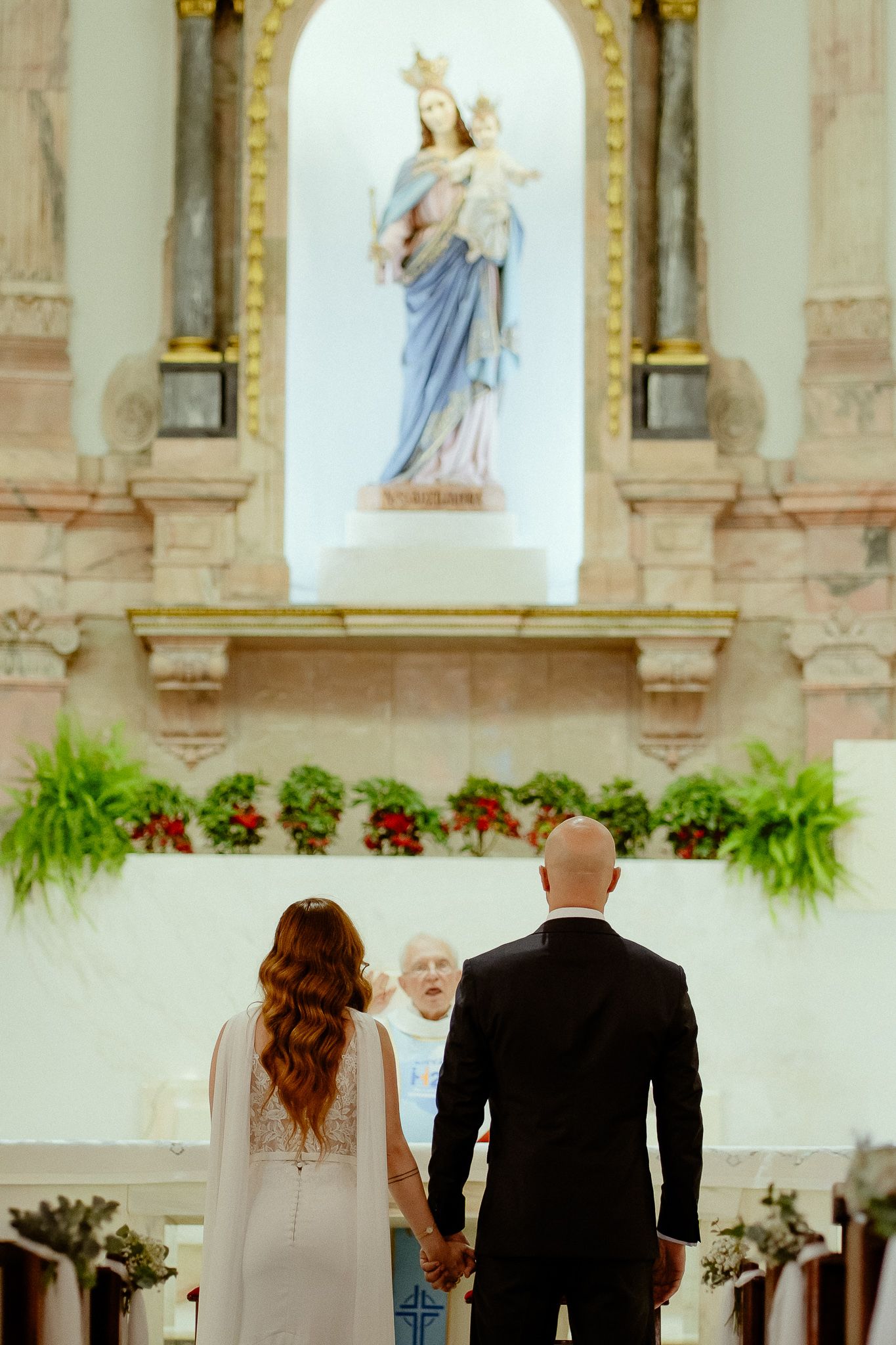 Bride and groom holding hands at church altar with Virgin Mary statue and officiant during wedding ceremony.
