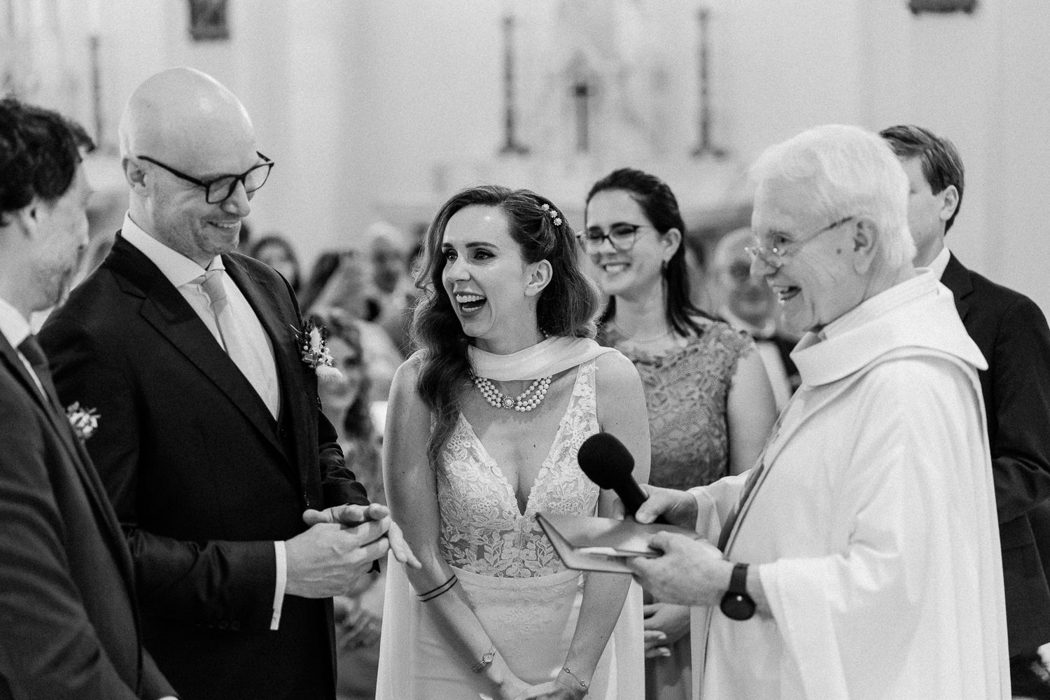Bride and groom laughing joyfully during wedding ceremony with priest officiating in elegant church.