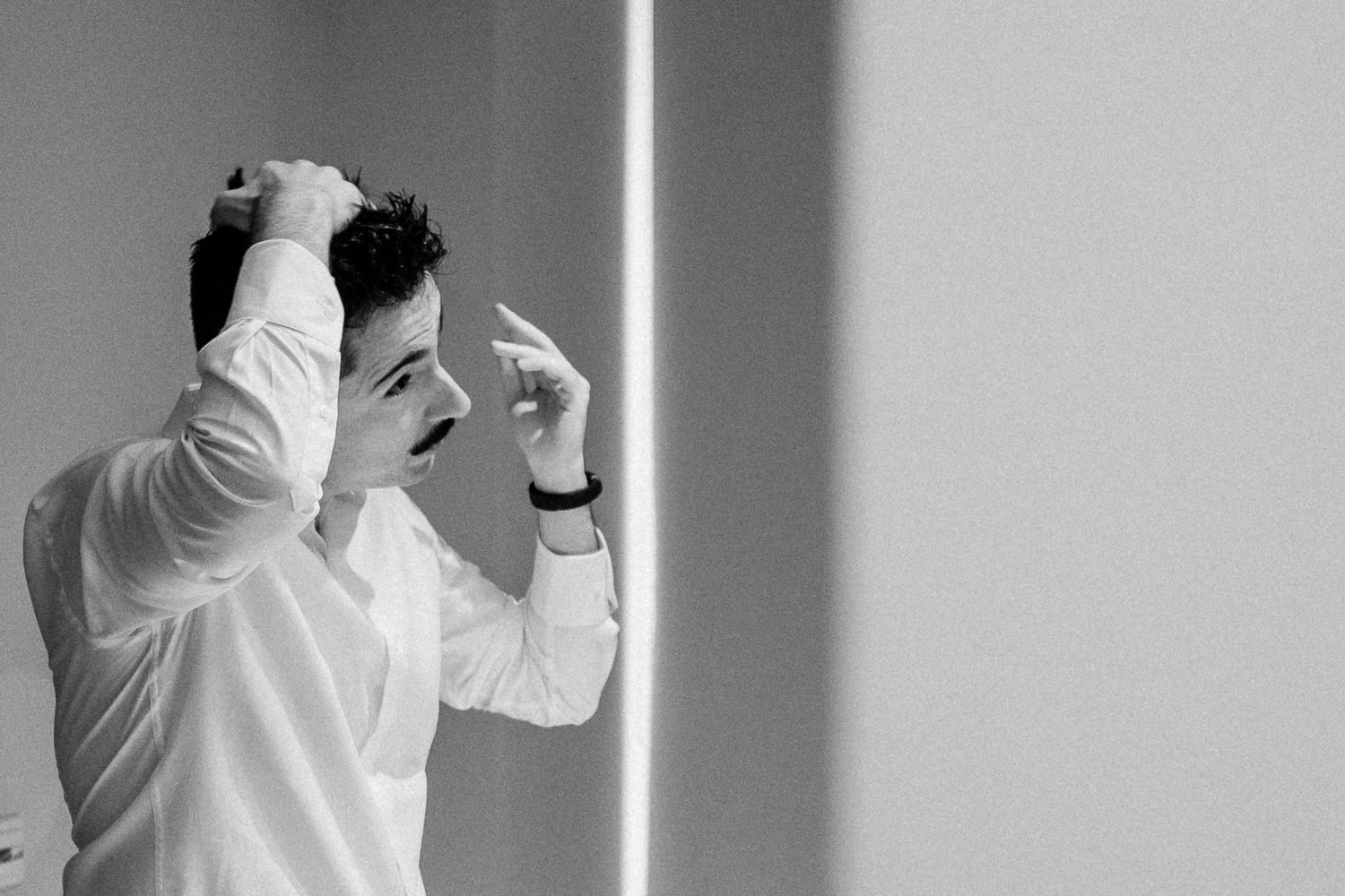 Black and white photo of a stressed man in white shirt holding his head with both hands against a light wall.