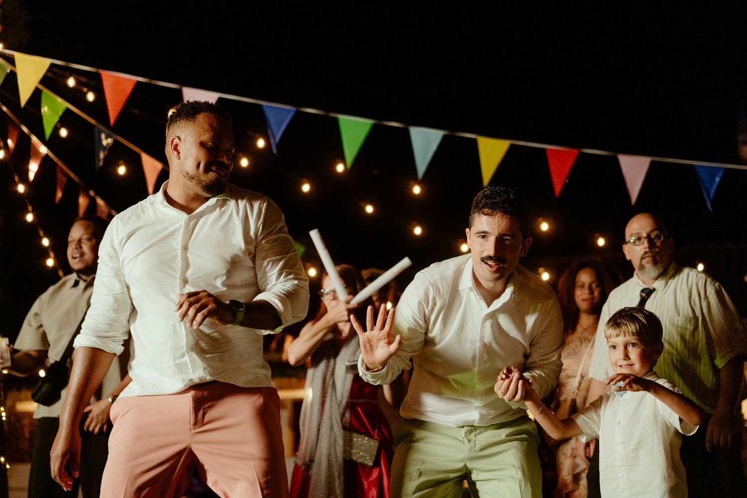 Two men in white shirts dance at a festive night party with colorful bunting and string lights.