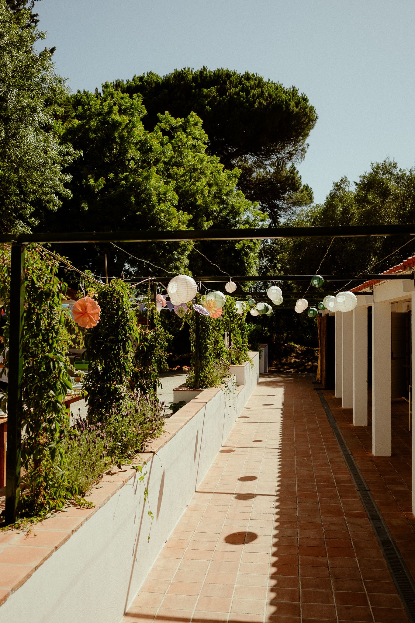 Outdoor corridor with colorful hanging lanterns, lush green trees, and terracotta tile flooring on a sunny day.