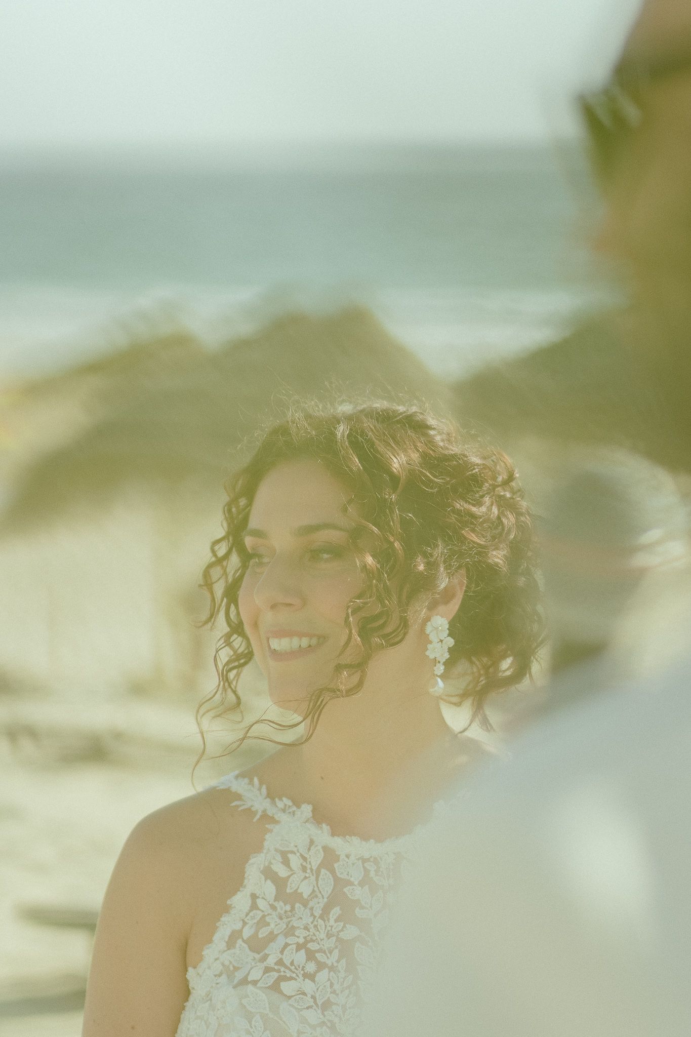 Smiling bride with curly hair and lace wedding dress stands on a scenic beach, with ocean waves visible in the background.