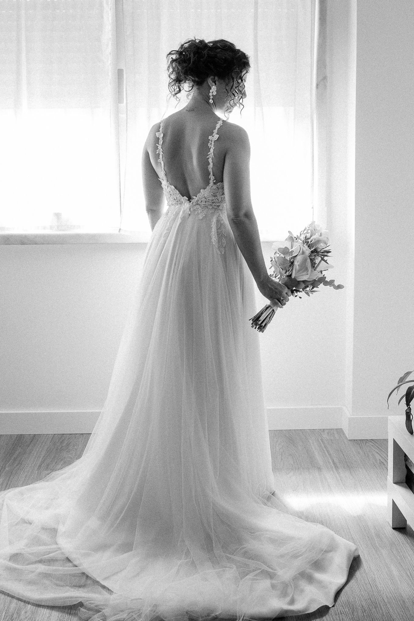 Elegant bride in backless lace wedding gown holding bouquet, standing by window in black and white photo.
