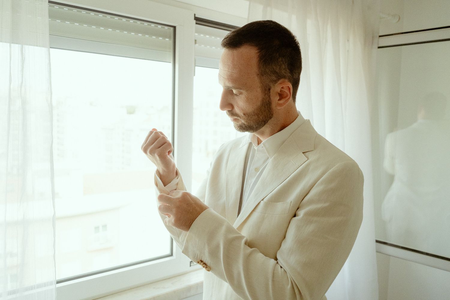 Man in white suit adjusting cufflinks near bright window, elegant wedding or formal event preparation.