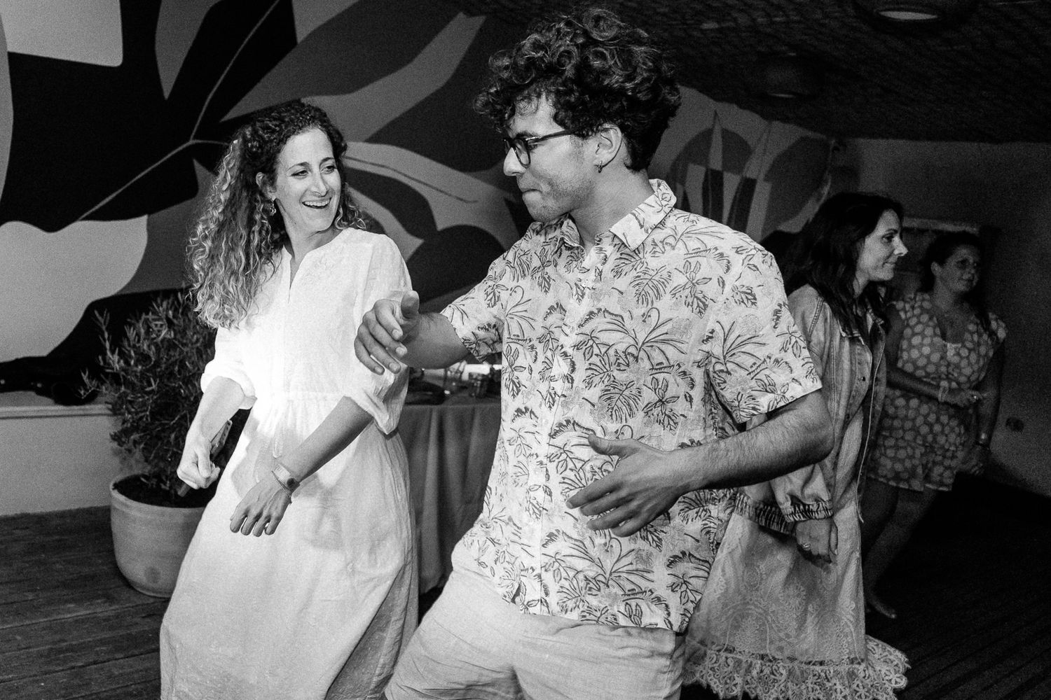 Two people dancing joyfully at a party in black and white photo, woman in white dress and man in floral shirt.