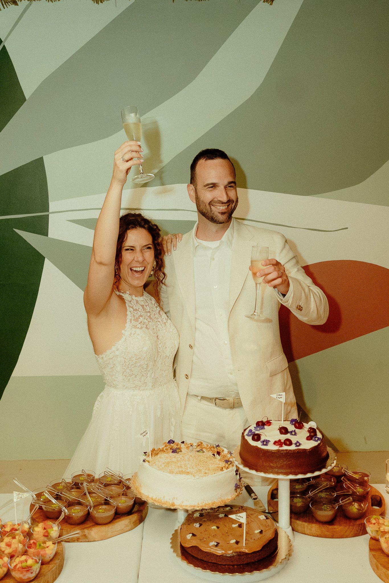 Bride and groom toasting with champagne glasses over a dessert table with wedding cakes at their reception.