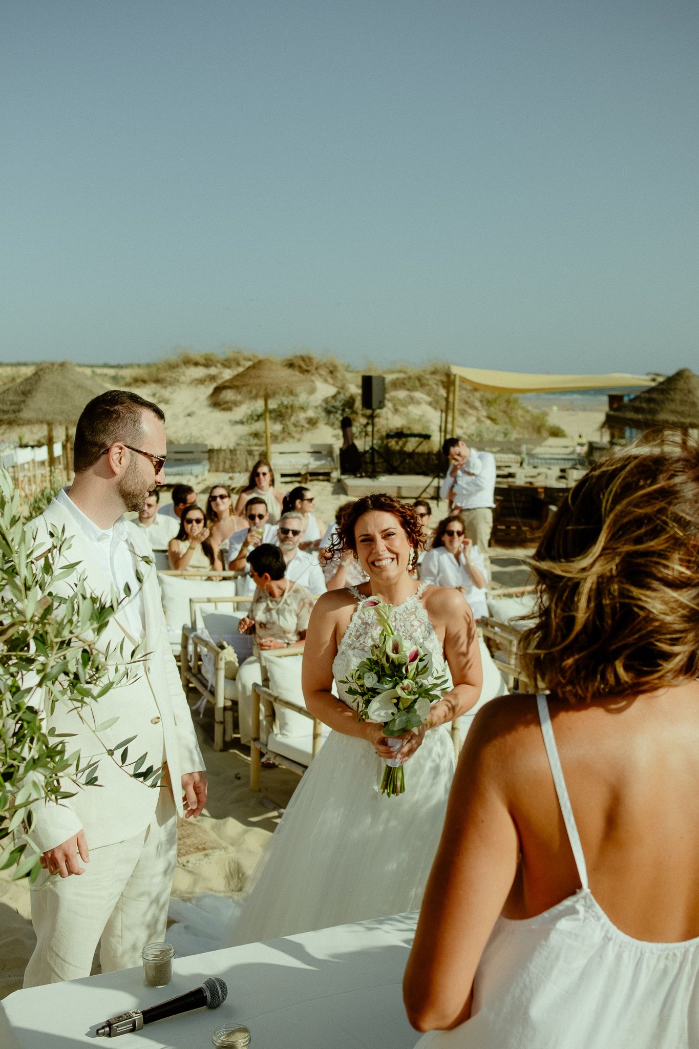 Bride smiling holding bouquet at outdoor beach wedding ceremony with groom in white suit and seated guests.