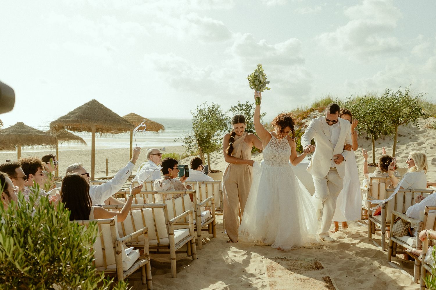 Bride and groom celebrate beach wedding ceremony with guests seated on sandy shore under thatched umbrellas.
