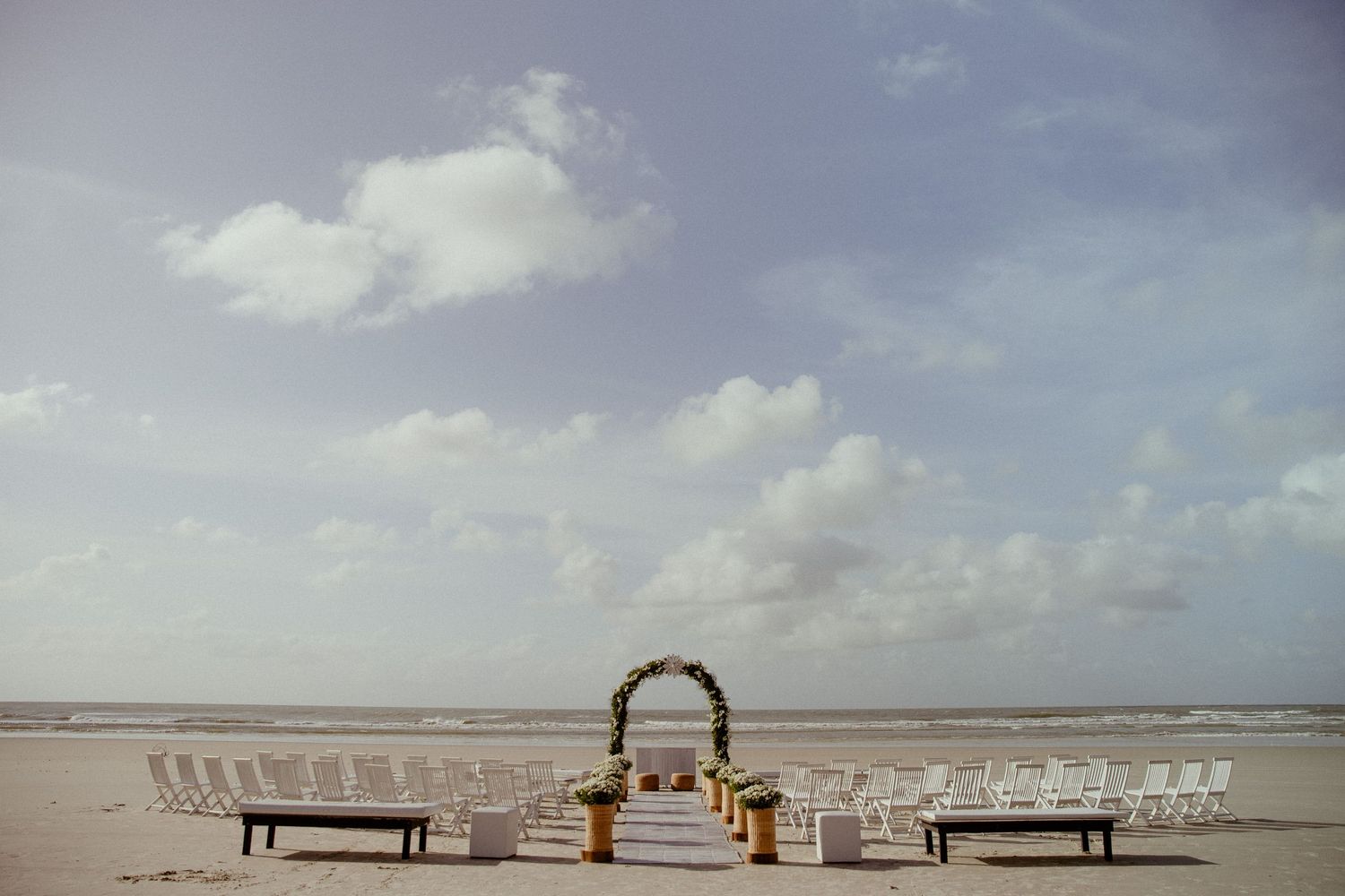 Wedding ceremony setup with white chairs and wooden arch on empty beach under cloudy sky at sunset.