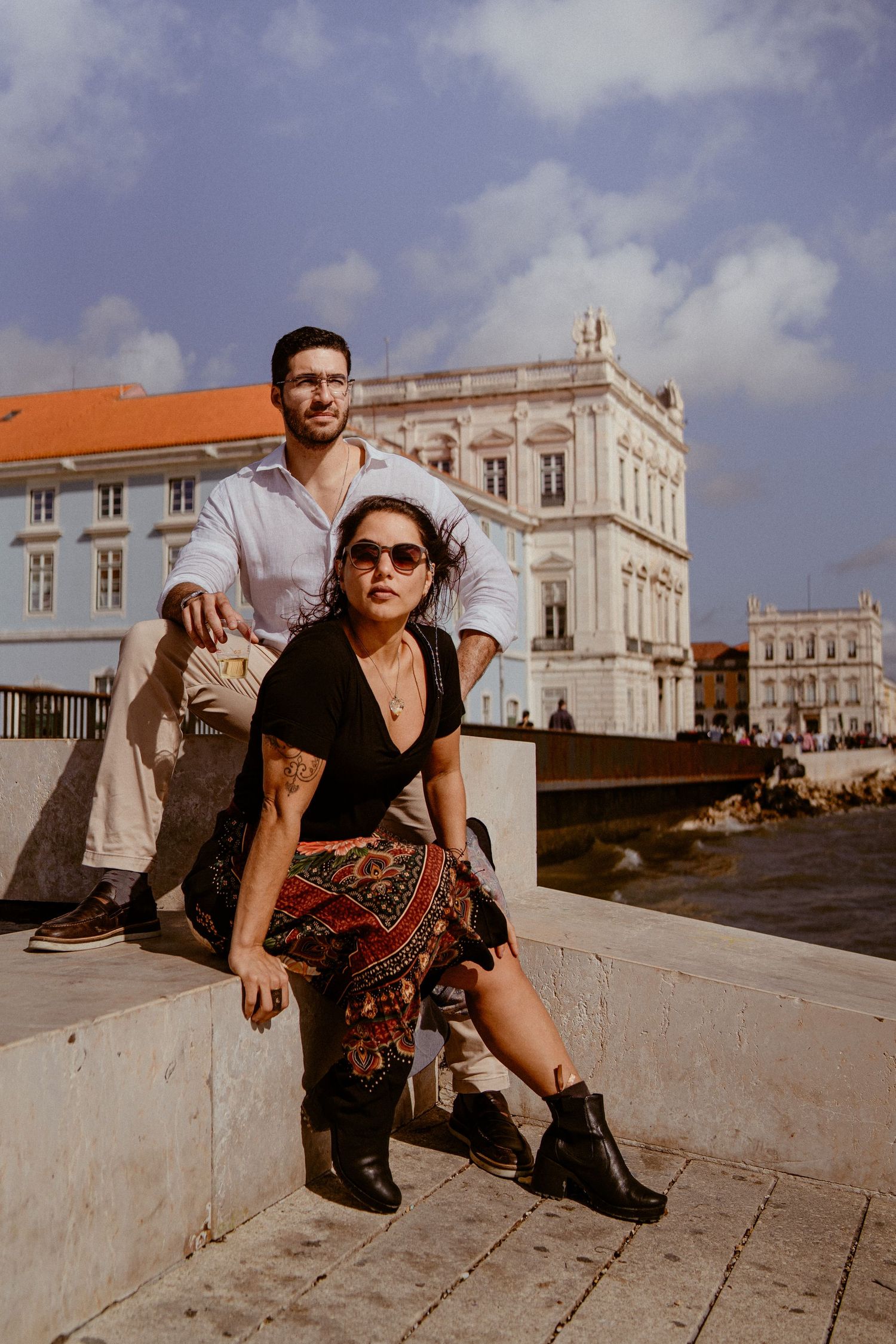 Fashion model poses against white stone wall with historic European building facade visible in warm sunlight.