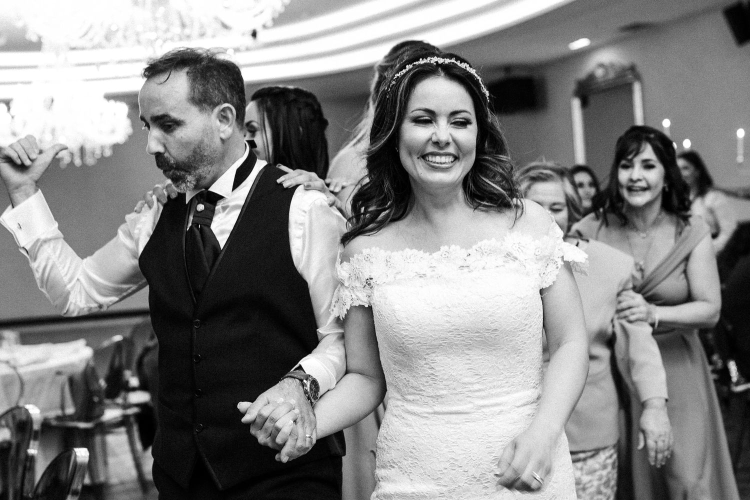 A newlywed couple walks hand in hand during their reception celebration while guests follow behind in black and white.