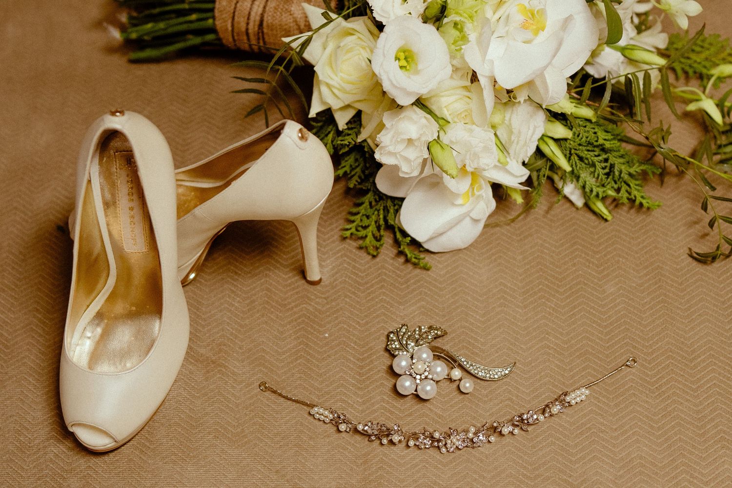 Elegant wedding accessories including white heels, pearl jewelry, and floral arrangements on beige fabric.