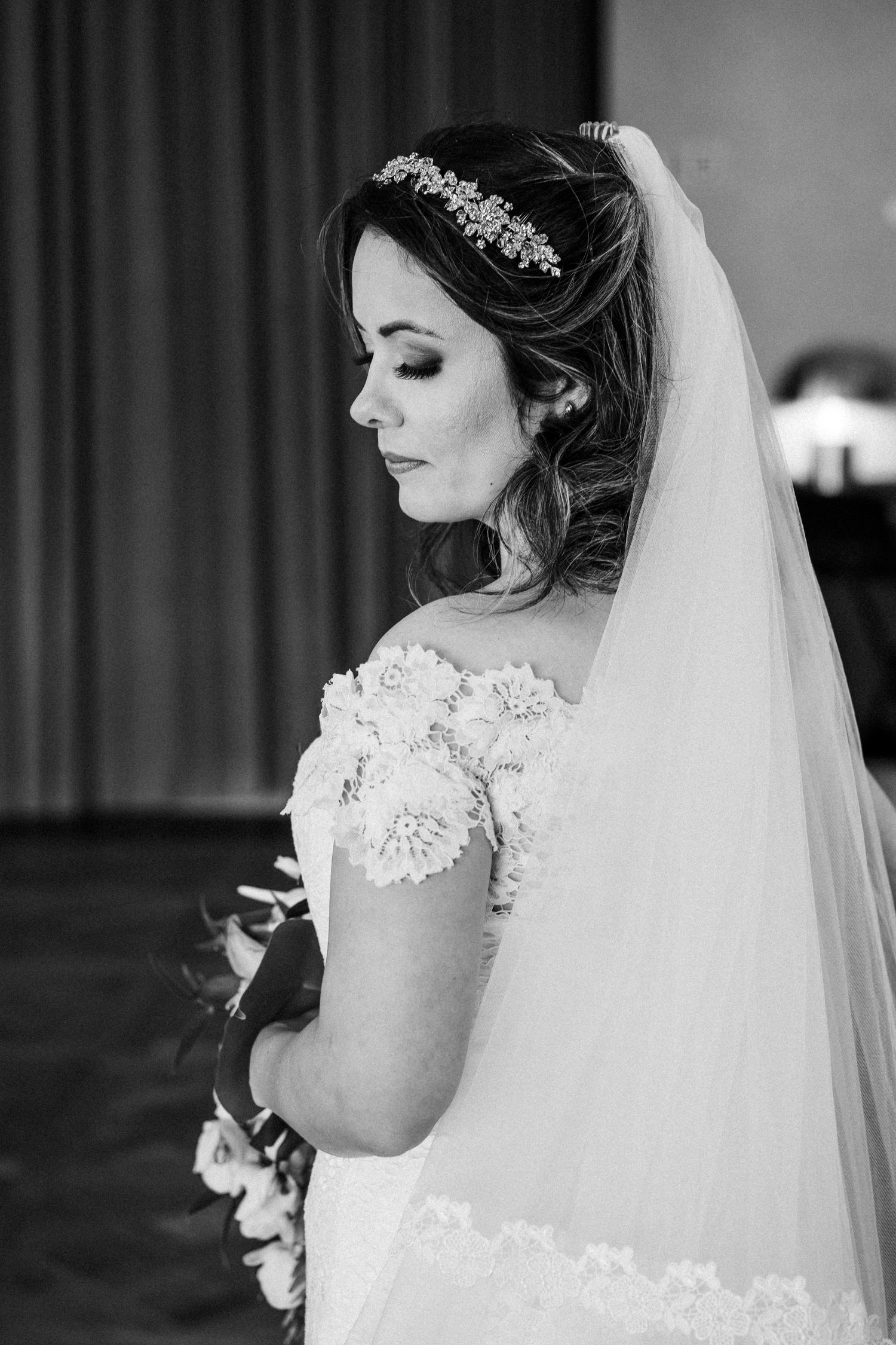 Elegant bride in lace gown and crystal headpiece poses with flowing veil in black and white portrait.