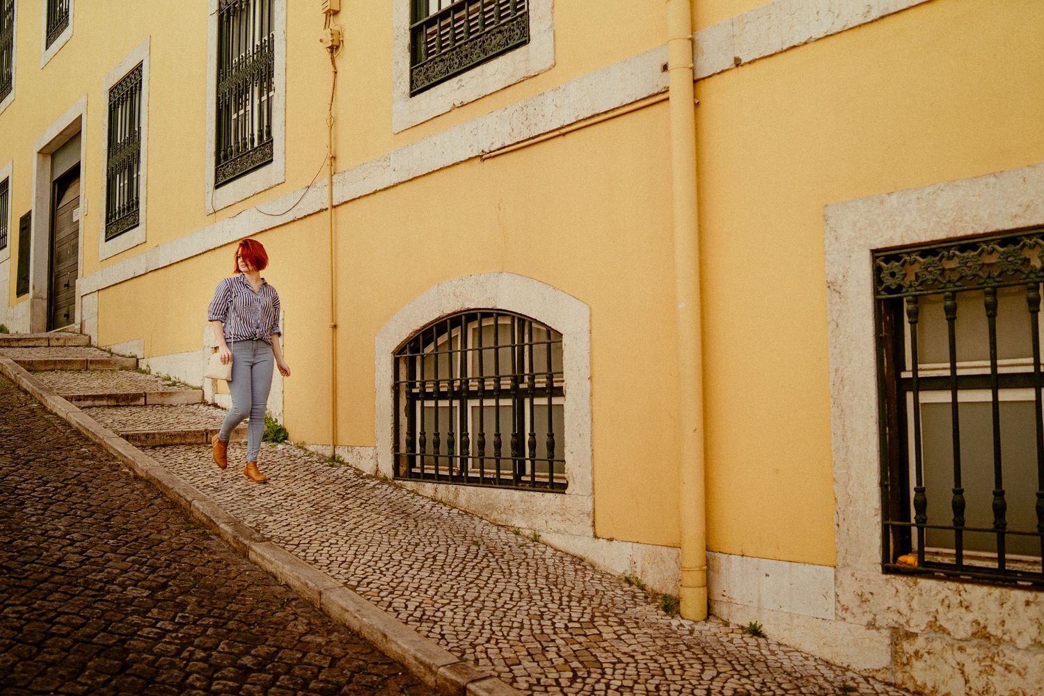 Person in purple shirt walking up cobblestone street by yellow Mediterranean building.