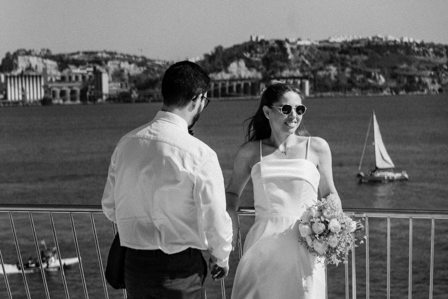 Black and white photo of a bride in sunglasses holding a bouquet facing groom by waterfront with sailboat backdrop.