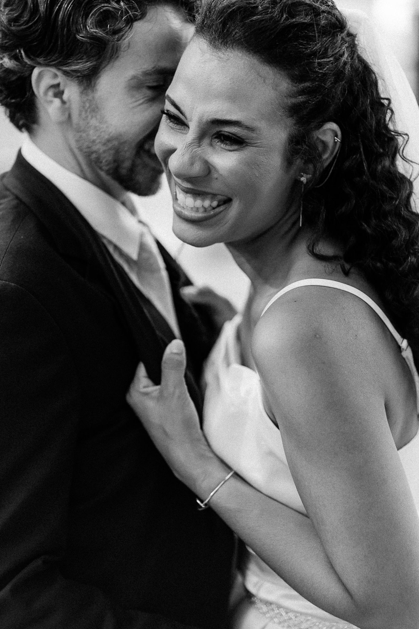 Black and white photo of a joyful bride smiling at groom in suit, sharing an intimate moment at their wedding.