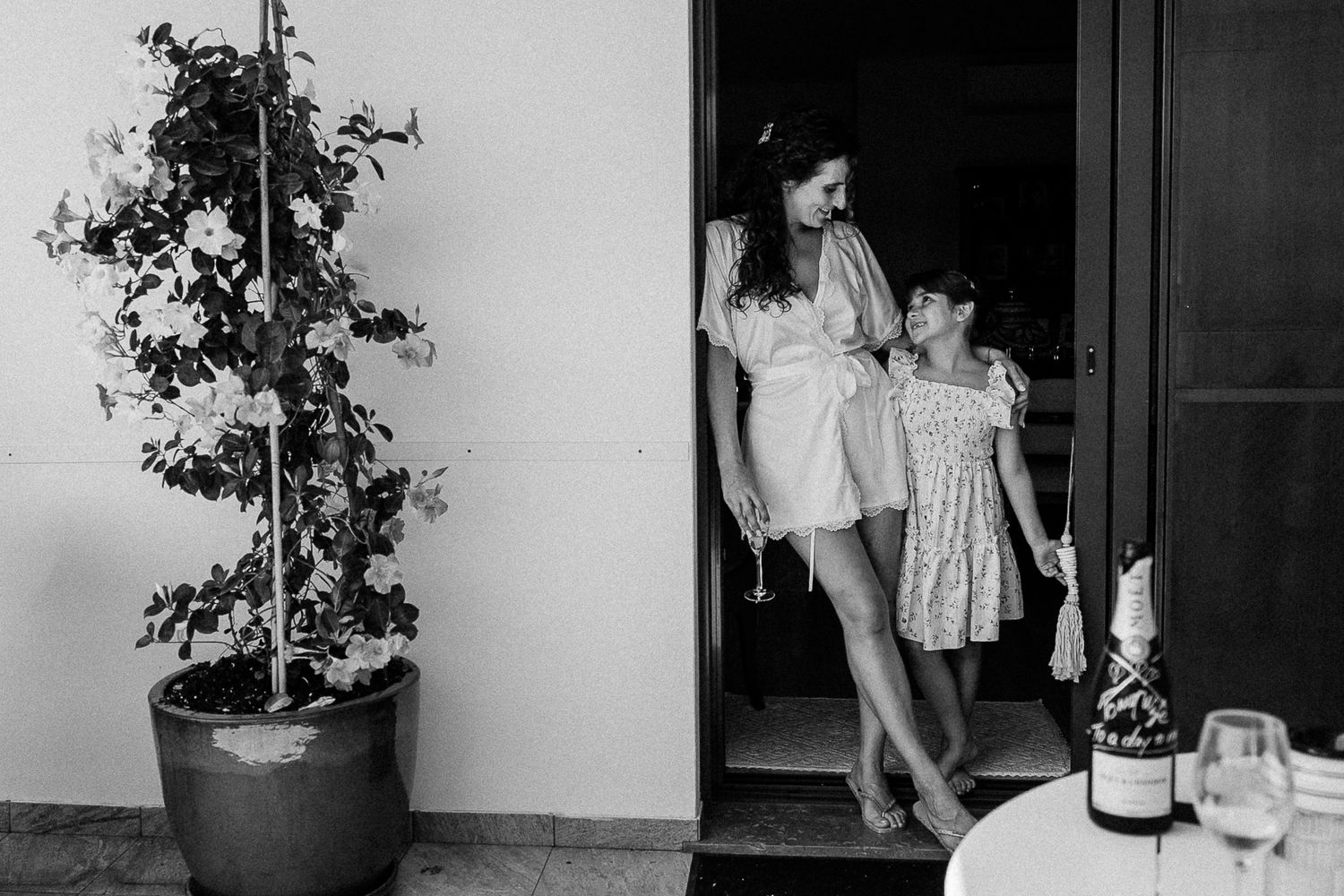 Black and white photo of two women embracing in a doorway beside a potted plant and a table with wine glasses.