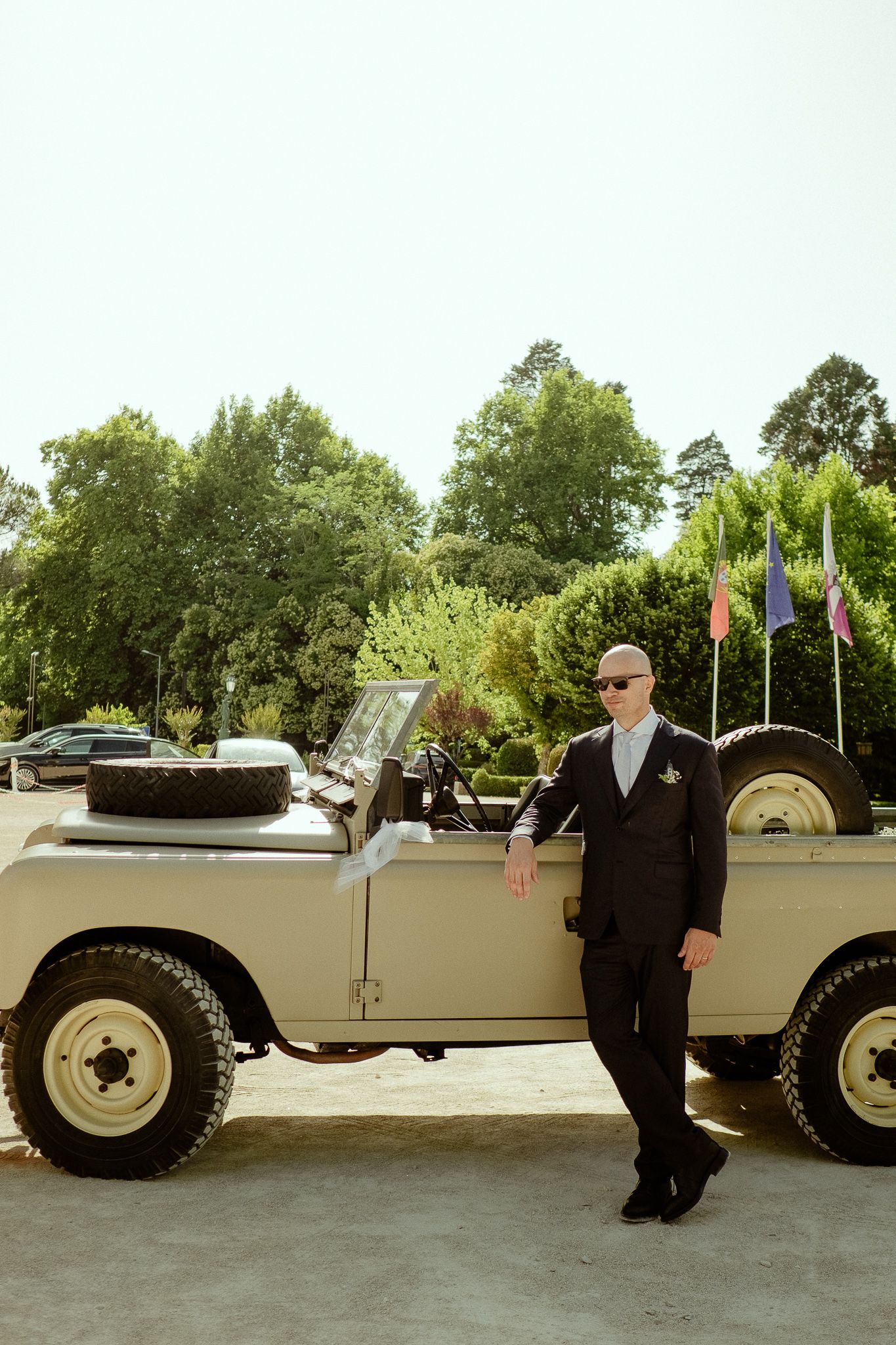 Bald man in suit leaning against vintage cream Land Rover with flags and trees in background.