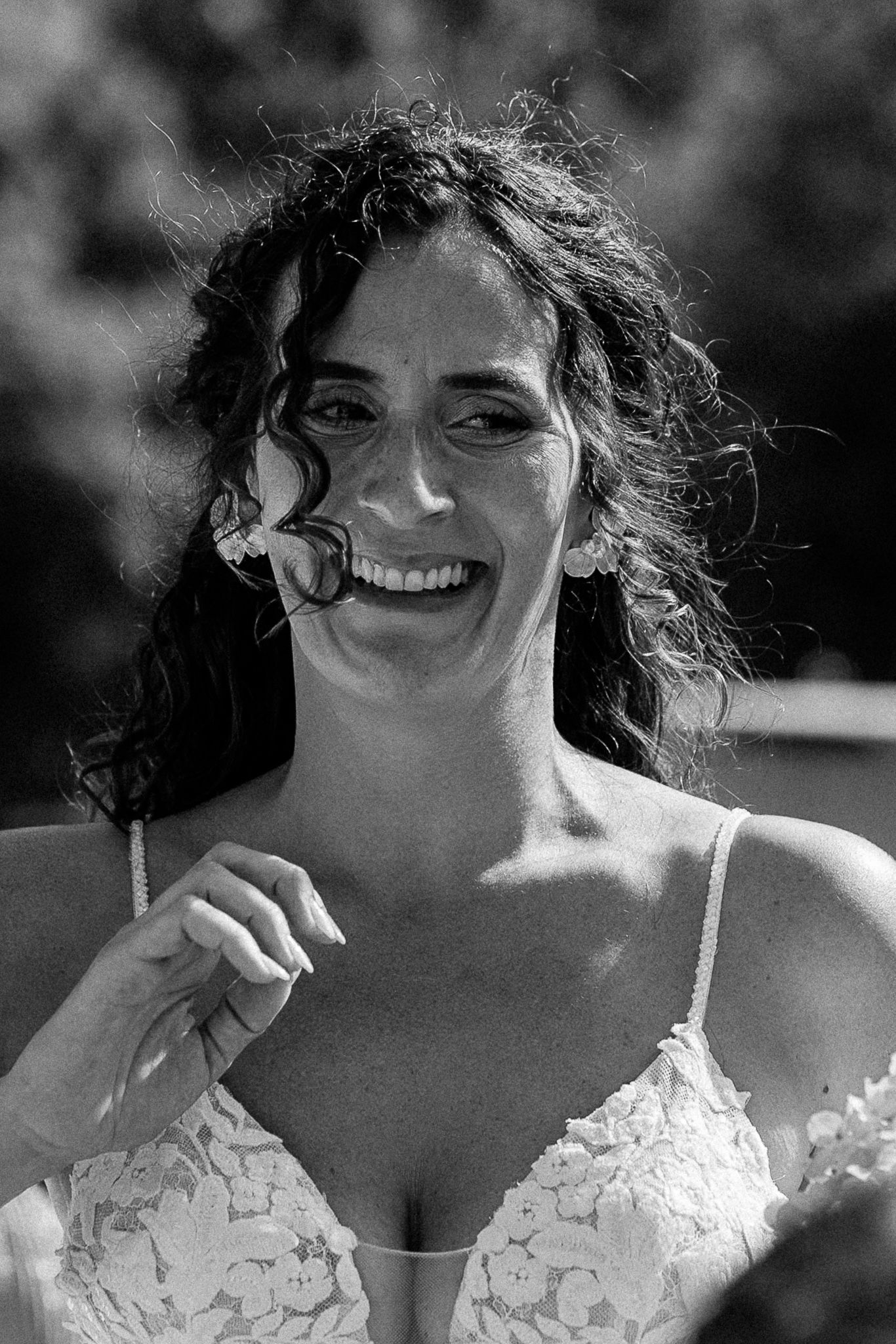 Black and white portrait of a smiling bride in lace dress, curly hair blowing in the breeze outdoors.