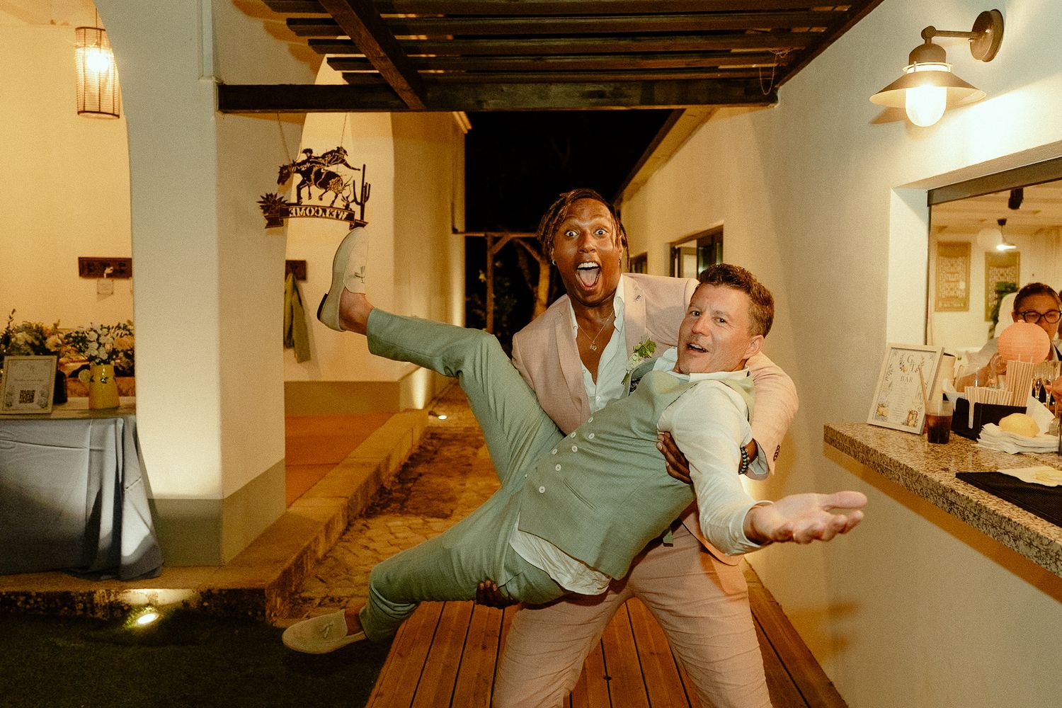 Two men celebrating joyfully at a wedding reception, one lifting the other in a fun pose indoors.