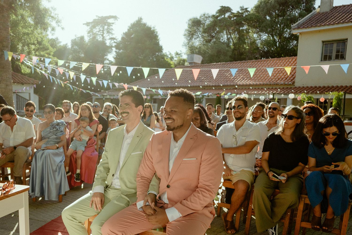 Two men in colorful suits smile during an outdoor wedding ceremony with festive bunting and seated guests.