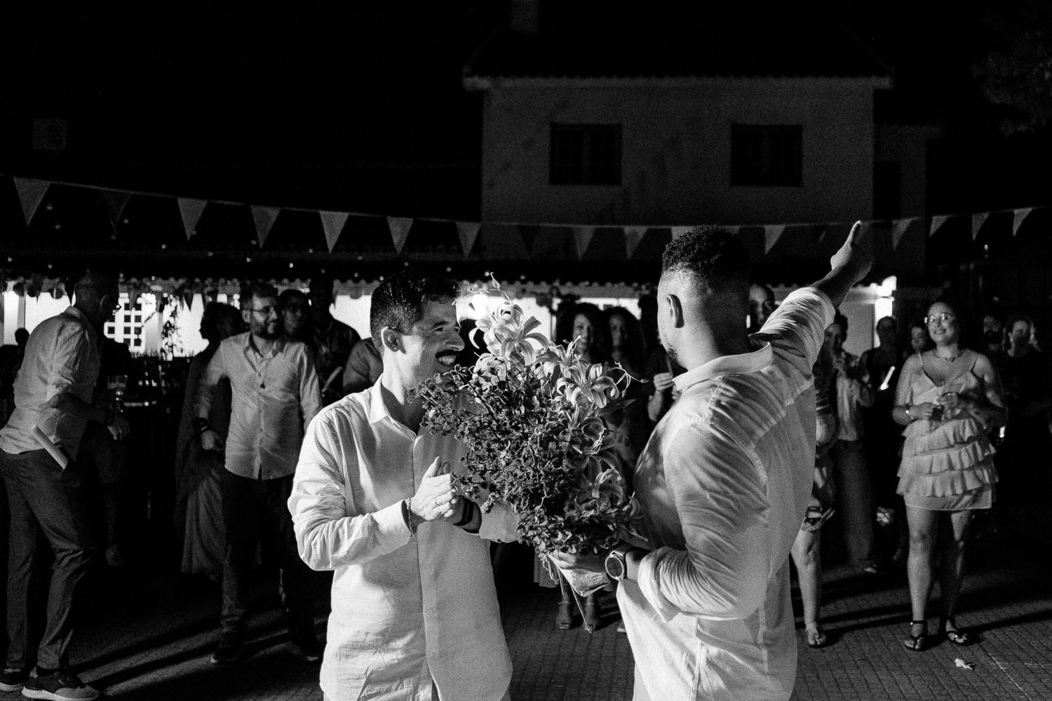 Two grooms share a romantic moment with flowers at an outdoor evening wedding reception.